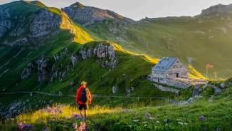 Un escursionista guarda il rifugio Pfälzerhütte nel Liechtenstein, circondato da un verde paesaggio montano e da un panorama alpino nella luce della sera