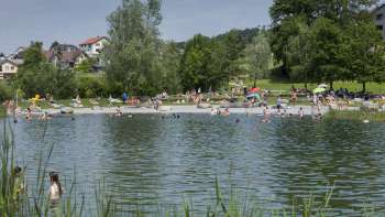 Giornata di bagni estivi nel laghetto naturale dell'Impianto ricreativo di Grossabünt, circondato da prati verdi e alberi.
