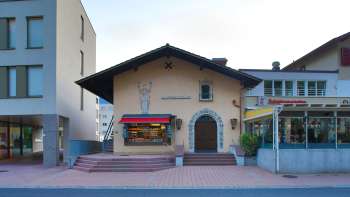 Vista esterna del vecchio cinema di Vaduz con le scritte sopra l'ingresso e le tende da sole rosse, situato tra due edifici moderni.