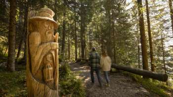 Due escursionisti guardano le figure di legno intagliate sul «sentiero delle leggende Walser» un percorso tematico nel mezzo di un paesaggio alpino con vista sulle montagne