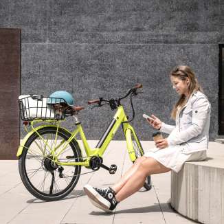 Giovane donna seduta con caffè e smartphone accanto a una e-bike gialla nel centro urbano di Vaduz