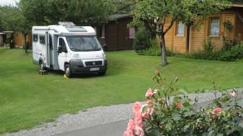 Piazzola per camper nel campeggio Mittagsspitze con prato verde e capanne in legno circostanti.