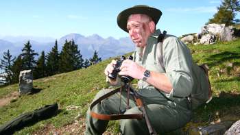Un cacciatore con un binocolo siede in un prato sullo sfondo di una montagna alpina.