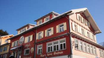 Vista esterna dell'Alpenhotel Vögeli Malbun con l'Elchbar al piano terra - edificio tradizionale rosso con scritte bianche contro un cielo blu.