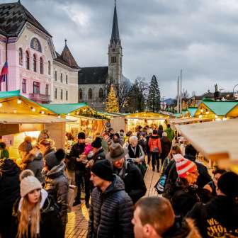 Atmosfera serale del mercatino di Natale di Vaduz 2018 con bancarelle addobbate a festa e folla di persone di fronte al palazzo del governo e alla cattedrale