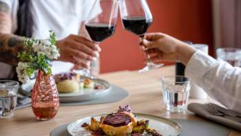 Due persone brindano con vino rosso, in primo piano un piatto principale preparato con cura