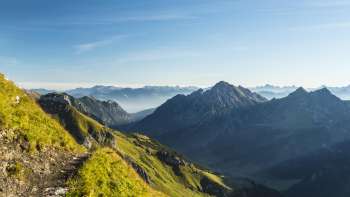 Sentiero escursionistico alpino con vista su un paesaggio montano verdeggiante e cime aspre al sole del mattino