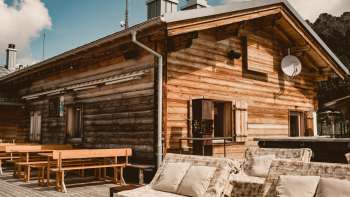 Accogliente terrazza solarium con panche in legno e comodi mobili da salotto di fronte al rifugio Sareis, a 2.000 metri di altitudine, con un sole splendente e lo sfondo delle montagne.