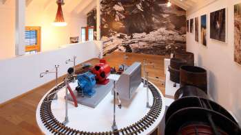 Modello di turbina su una piattaforma espositiva del Museo Lawena, con fotografie storiche in bianco e nero sullo sfondo.