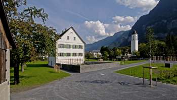 Canonica storica con facciata bianca e persiane verdi al centro di una piazza aperta, con la chiesa di Balzers e il paesaggio montano circostante sullo sfondo.