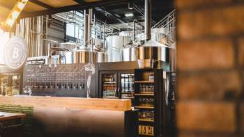 Vista interna di una birreria con impianto di produzione in acciaio inox e invitante bar in primo piano.
