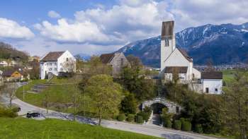 Vista panoramica della «Kirchhügel» (collina della chiesa) di Bendern con la sua chiesa storica, circondata da sentieri curati, alberi e architettura tradizionale sullo sfondo delle montagne alpine.