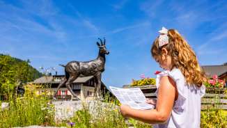 Ragazza con mappa davanti alla statua del camoscio a Malbun - tour esplorativo a misura di bambino nella località turistica per famiglie del Liechtenstein.