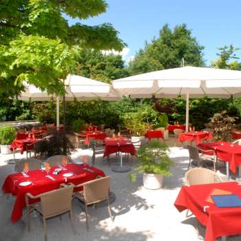Vista esterna della terrazza con tovaglie rosse, circondata da alberi e ombrelloni.