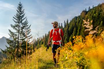 Escursionista in maglietta rossa su un soleggiato sentiero di montagna vicino a Malbun, circondato da fiori di prato, boschi di conifere e paesaggi alpini nel Liechtenstein