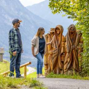 Una coppia osserva le figure di legno intagliate sul Sentiero della leggenda Walser a Triesenberg con vista sulla valle e sulle montagne sullo sfondo
