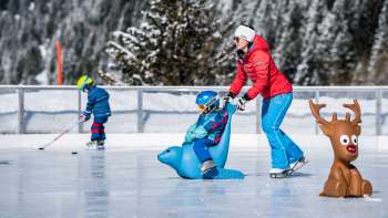 Bambini sulla pista di ghiaccio punto di ritrovo Schlucher-Treff a Malbun, sostenuti da un ausilio per il pattinaggio a forma di animale blu.