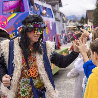 Hippie travestito al carnevale dei bambini di Mauren, dà il cinque ai bambini davanti a un carro colorato.