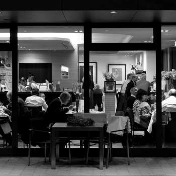 Foto in bianco e nero di un ristorante affollato con vista dall'esterno attraverso la facciata in vetro.