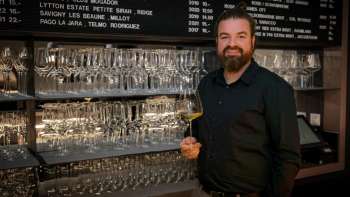 Barista amichevole dell'WY Weinbar con bicchiere di vino davanti a un elegante scaffale pieno di bicchieri e vini.