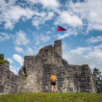 Escursionista davanti alle rovine storiche del castello di Schellenberg con la bandiera del Liechtenstein che sventola sotto un cielo azzurro