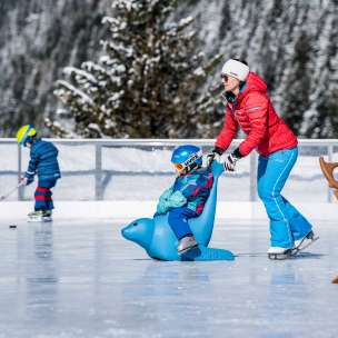 Madre spinge il figlio su un pinguino che pattina sul ghiaccio attraverso la pista di Malbun, sullo sfondo un bambino con una mazza da hockey su ghiaccio