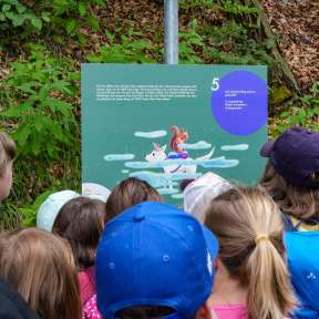 Gruppo di bambini osserva un pannello informativo con un'illustrazione di "Mila nel tunnel delle meraviglie" lungo il sentiero di Gafadura nella foresta.