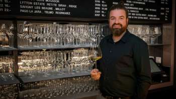 Barista amichevole dell'WY Weinbar con bicchiere di vino davanti a un elegante scaffale pieno di bicchieri e vini.