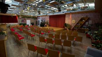 Ampia e luminosa sala per eventi della Kreativ Akademie con palco, tende rosse e file di sedie - ideale per seminari, conferenze ed eventi creativi in Liechtenstein.