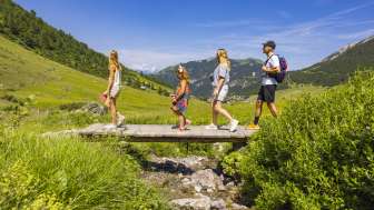 Una famiglia attraversa un piccolo ponte di legno durante un'escursione nel paesaggio alpino di Malbun, in Liechtenstein, sotto un sole splendente.