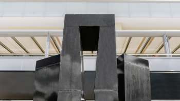 Scultura "Progressione di una forma in 3 stele" di Christian Megert in pietra nera davanti a una moderna facciata di vetro a Vaduz.