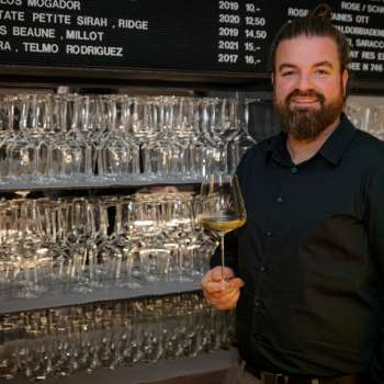 Barista amichevole dell'WY Weinbar con bicchiere di vino davanti a un elegante scaffale pieno di bicchieri e vini.
