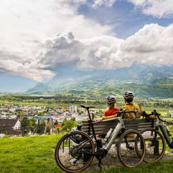 Due ciclisti con il casco si siedono su una panchina sopra la valle del Reno e si godono la vista della valle e delle montagne circostanti - un'esperienza di gravel bike in Liechtenstein