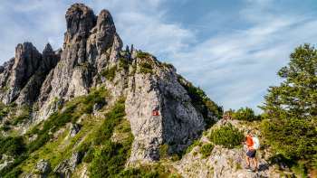 Escursionisti su un sentiero roccioso di montagna davanti a una cresta impervia con un cartello rosso e bianco