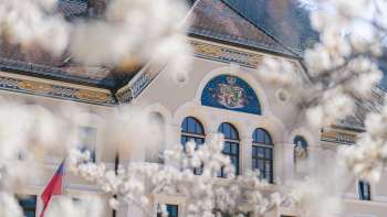 Il palazzo del governo di Vaduz incorniciato da alberi in fiore in primavera, in primo piano lo stemma nazionale.