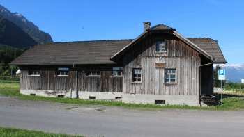 Lo storico edificio in legno della vecchia segheria di Schaanwald sullo sfondo alpino, sotto un cielo azzurro.