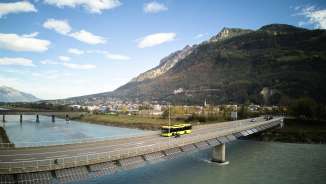 L'autobus giallo di LIEmobil attraversa il ponte sul Reno vicino a Vaduz con un panorama montano sullo sfondo - Experience Liechtenstein