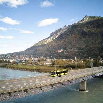 L'autobus giallo di LIEmobil attraversa il ponte sul Reno vicino a Vaduz con un panorama montano sullo sfondo - Experience Liechtenstein