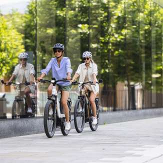 Due donne in e-bike attraversano il moderno centro di Vaduz, in Liechtenstein, con la sua facciata di vetro e il suo stile urbano.