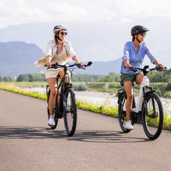Due donne in e-bike sulla pista ciclabile del Reno, vicino al Liechtenstein, con il fiume, il paesaggio verde e le montagne sullo sfondo