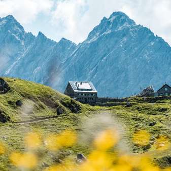 Vista lontana del rifugio Pfälzerhütte nel Liechtenstein con fiori selvatici in primo piano e ripide cime rocciose sullo sfondo