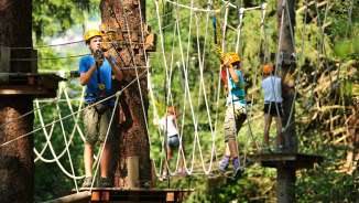Bambini e giovani con il casco nel percorso a corde alte del parco di arrampicata su corde di Triesen in Liechtenstein, assicurati agli elementi di arrampicata nella foresta