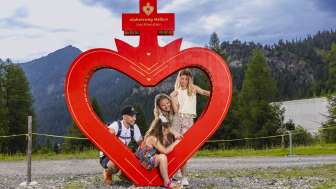 La famiglia posa davanti alla cornice del cuore rosso lungo il sentiero del cuore alpino di Malbun, nel Liechtenstein, circondata dalla natura alpina e dalle montagne.