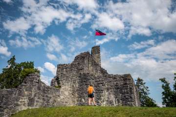 Escursionista davanti alle rovine storiche del castello di Schellenberg con la bandiera del Liechtenstein che sventola sotto un cielo azzurro