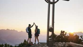 Due escursionisti si danno il cinque alla croce di vetta dell'Alpspitz all'alba, con un panorama montano sullo sfondo.