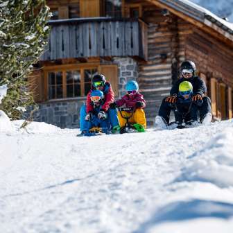 Due adulti e tre bambini siedono sulle slitte e iniziano una discesa insieme davanti a una baita innevata a Malbun