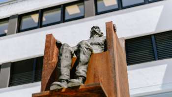 Scultura "Hochsitz" di Robert Indermaur: una figura è seduta su una sedia d'acciaio color ruggine di fronte a una facciata moderna.