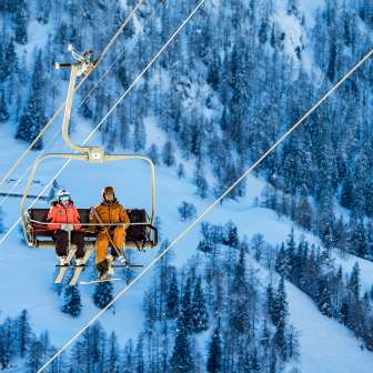 Tina Weirather e Marco Büchel siedono insieme su una seggiovia, circondati da alberi innevati e paesaggi invernali a Malbun.